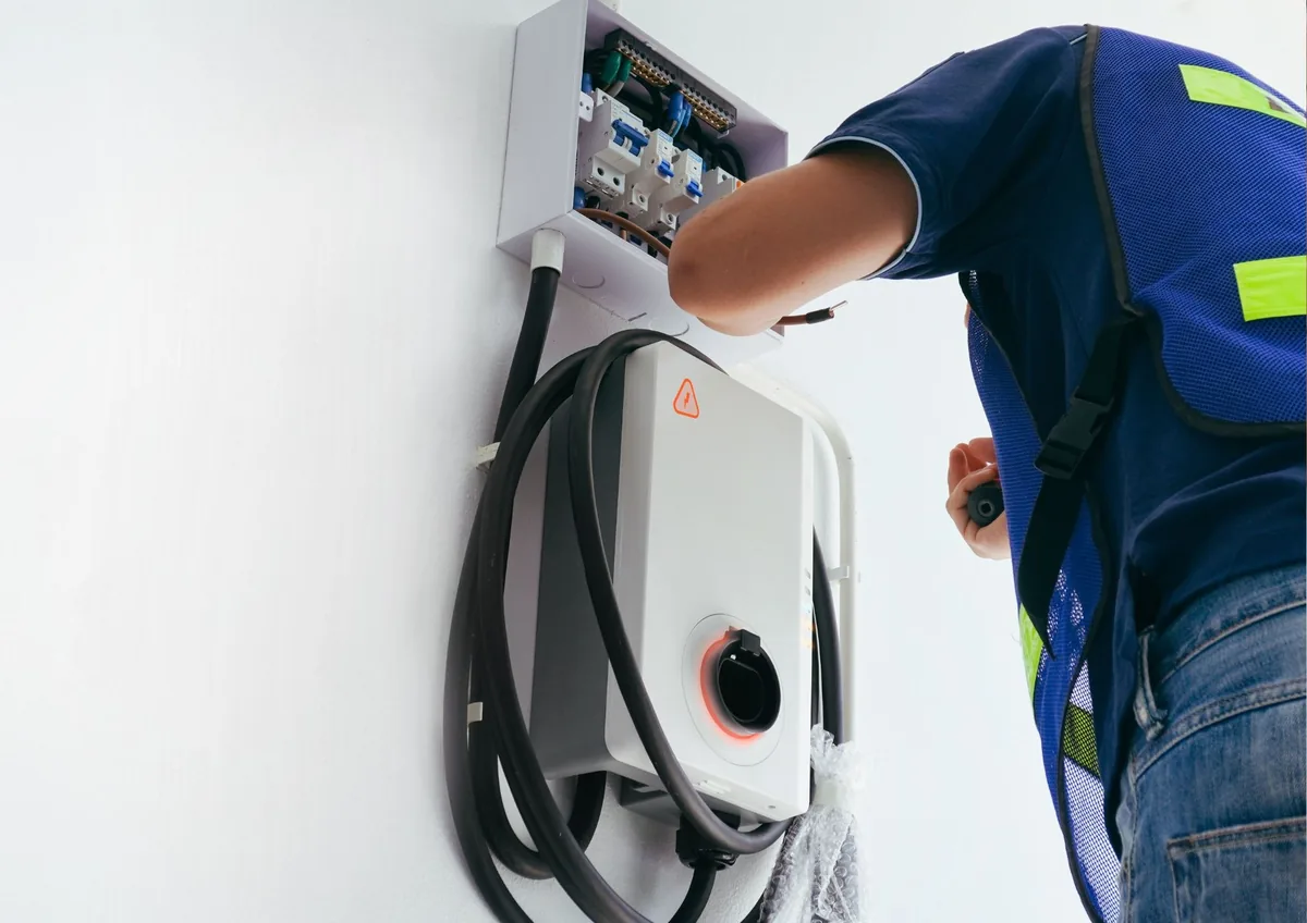 Electrician in a safety vest wiring a small breaker box above a wall-mounted EV charger with its cable coiled